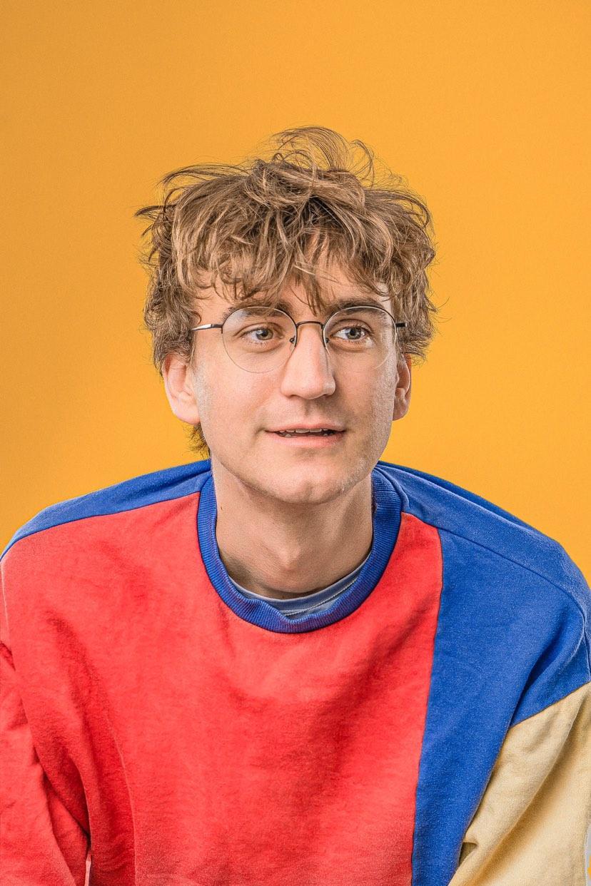 Image of Sam Coade, who has curly blonde hair and glasses and is wearing a colourful jumper